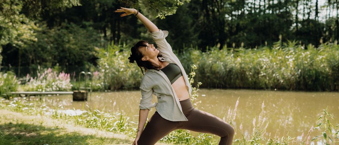 Eine Frau praktiziert Yoga auf einer Matte in einem Park mit einem ruhigen See und Bäumen im Hintergrund. Sie ist barfuß und streckt Arme und Beine.