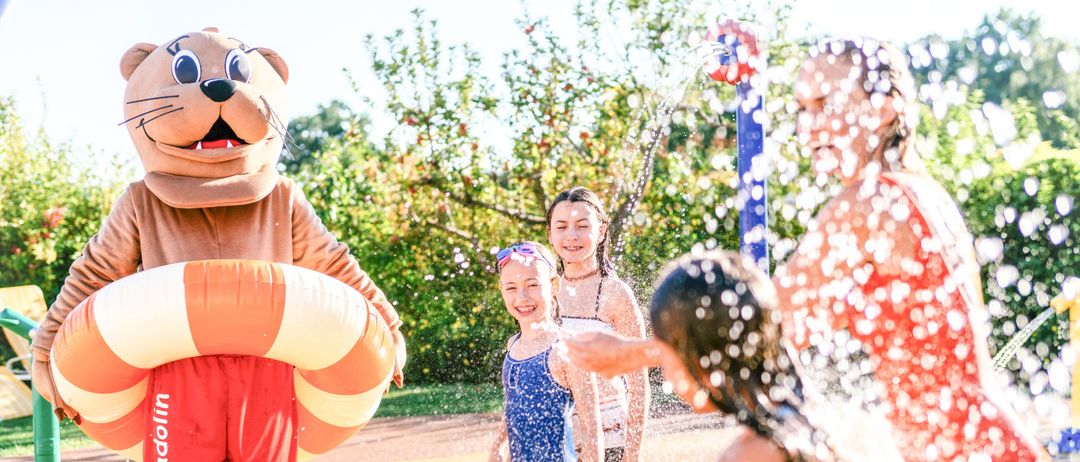 Ein Maskottchen in einem Bärenkostüm interagiert mit Kindern in einem Wasserpark, wobei Wasser um sie herum spritzt.