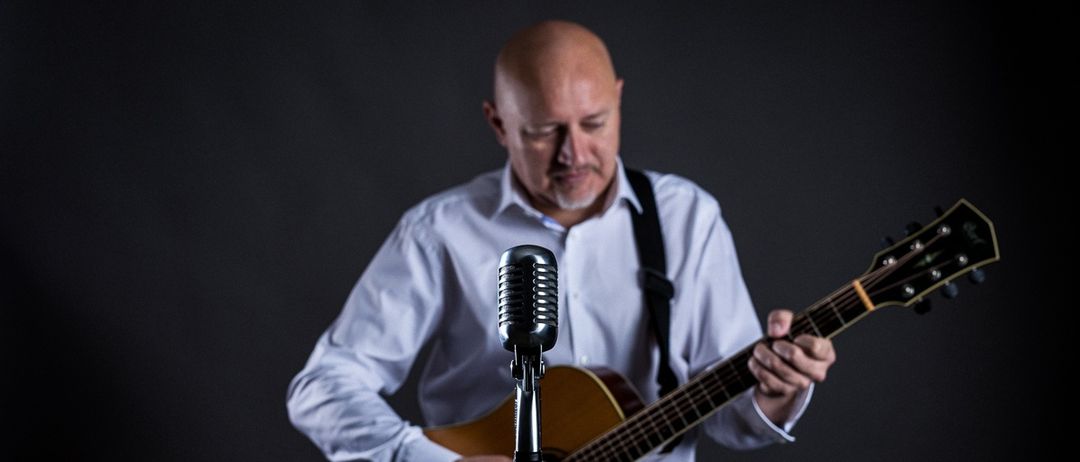 Ein kahlköpfiger Mann in einem weißen Hemd spielt Gitarre vor einem Mikrofon.