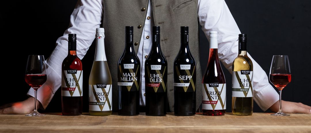 Ein Mann mit Weste und Brille lächelt und posiert mit mehreren Flaschen Wein auf einem Holztisch vor ihm.