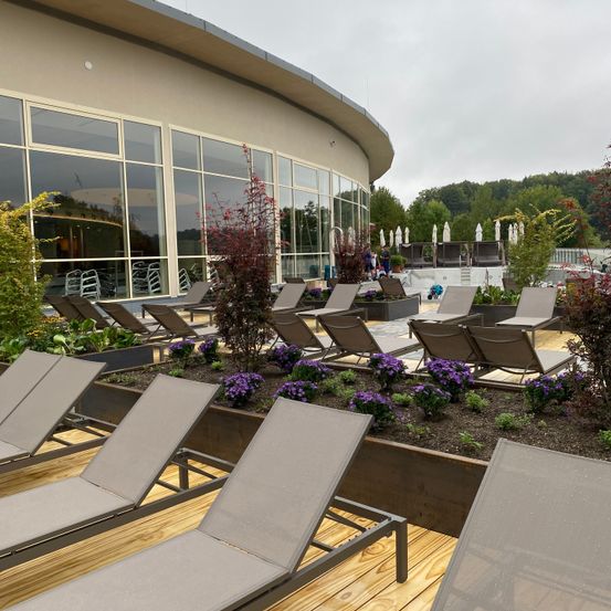 Freiluft-Loungebereich mit Holzdeck, grauen Liegestühlen und Pflanzen. Die Stühle sind in Reihen angeordnet, und es gibt Pflanzen dazwischen. Im Hintergrund befindet sich ein Gebäude mit Glasfenstern.