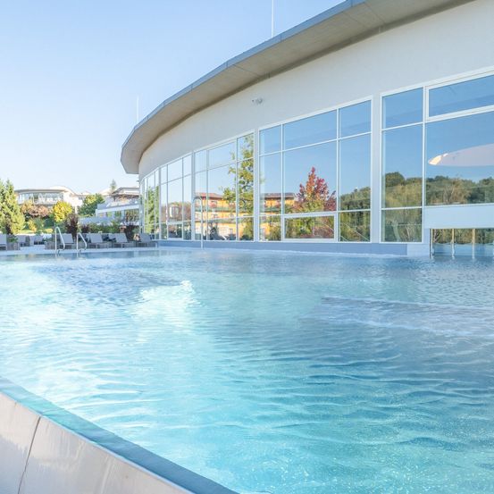 Ein Außenschwimmbad mit Glaswänden ist leer. Liegestühle sind auf der linken Seite aufgestellt.