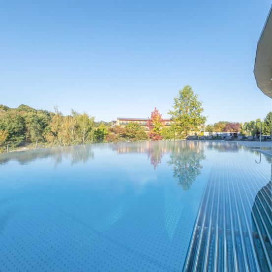 Ein großer Außenpool, umgeben von Bäumen und einem modernen Gebäude mit flachem Dach. Das Wasser ist klar und blau und reflektiert die Bäume und den Himmel.