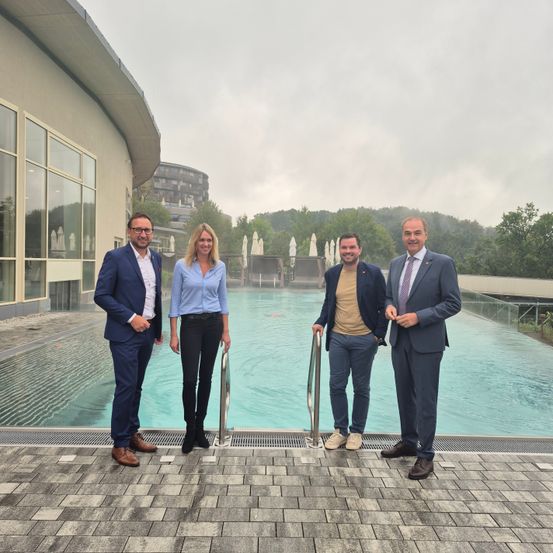 Vier Personen stehen in der Nähe eines großen Swimmingpools in einem Resort, mit einem modernen Gebäude im Hintergrund.