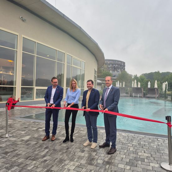 Vier Menschen in formeller Kleidung stehen neben einer roten Schleife, die den Eingang eines Schwimmbeckens versperrt. Hinter ihnen steht ein modernes Gebäude mit Glasfenstern und einem offenen Bereich mit Bäumen.