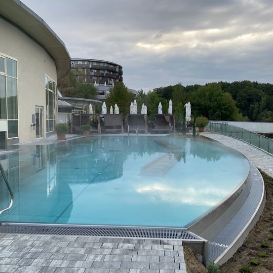 Ein moderner, geräumiger Außenpool, umgeben von einem Steinweg. Der Pool hat kristallklares blaues Wasser und um ihn herum befinden sich Liegestühle und geschlossene Sonnenschirme.