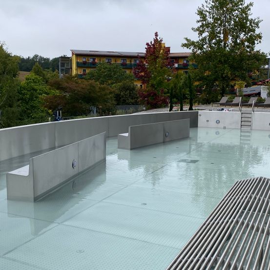 Ein Freibad umgeben von Bänken und Bäumen, mit einem gelben Gebäude im Hintergrund und einem bewölkten Himmel.