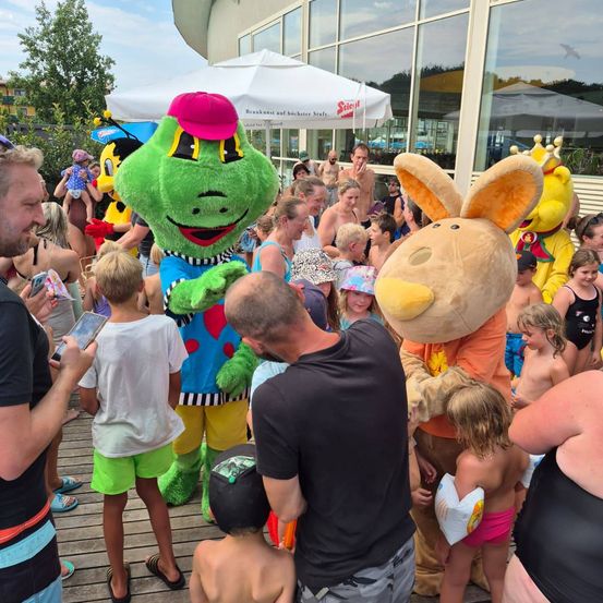 Eine Menschenmenge, darunter Kinder, versammelt sich um Plüschfiguren bei einer Veranstaltung im Freien. Eine der Plüschfiguren ist ein grüner Frosch mit einer rosa Mütze, und eine andere ist ein großer brauner Hase.