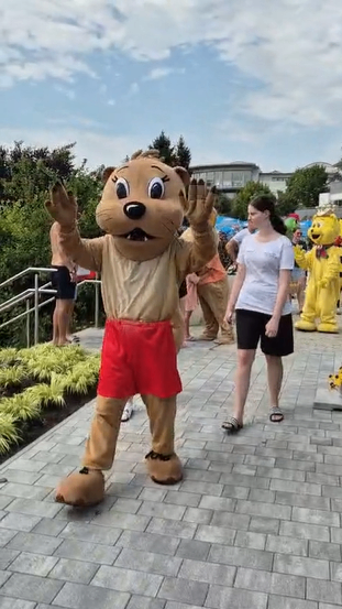 Eine Person in einem braunen Bären-Maskottchen-Kostüm winkt zur Kamera, während eine Frau in einem weißen T-Shirt in der Nähe auf einem Ziegelweg steht. Andere Maskottchen und Menschen sind im Hintergrund.