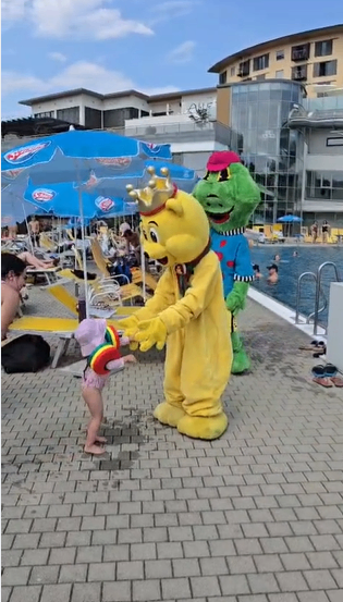 Ein gelber Maskottchen mit Krone interagiert mit einem kleinen Kind in der Nähe eines Schwimmbeckens, während ein grüner Maskottchen in der Nähe steht. Die Szene spielt in einem Resort mit Gebäuden und Menschen im Hintergrund.