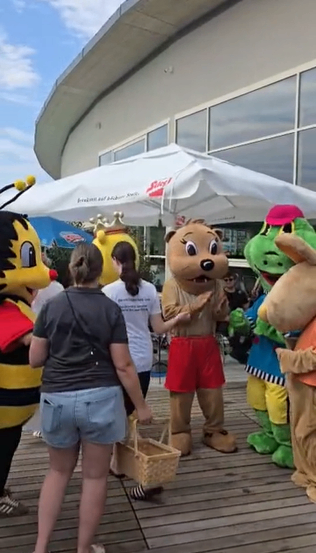 Eine Gruppe von Menschen, einschließlich einer Frau in Jeansshorts, steht in der Nähe von Maskottchen eines Bären, eines Frosches und einer Biene unter einem weißen Regenschirm. Die Maskottchen sind in Kostümen gekleidet und interagieren mit den Menschen.