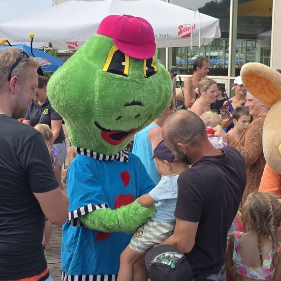 Ein Maskottchen in einem blauen Shirt mit roten Herzen interagiert mit einem kleinen Jungen und seinem Vater in einem überfüllten Außenbereich.