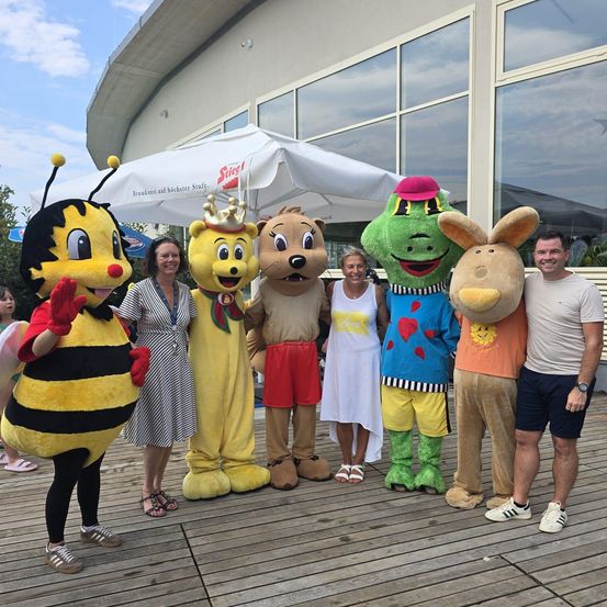 Eine Gruppe von Menschen und Maskottchen, darunter eine Biene, ein Bär, ein Frosch und ein Hase, posiert für ein Foto vor einem Gebäude.