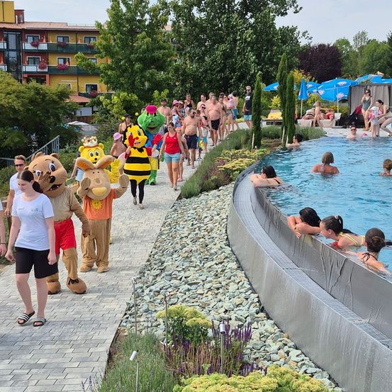 Eine Gruppe von Menschen, einige in Maskottchenkostümen, geht entlang eines Weges in der Nähe eines Schwimmbeckens. Das Becken ist mit Schwimmern gefüllt, und im Hintergrund befinden sich Schirme und Gebäude.
