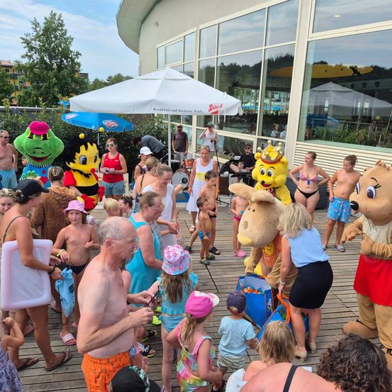 Eine Gruppe von Menschen, einschließlich Erwachsener und Kindern, versammelt sich um Plüschtiere auf einer Holzterrasse, einige in Badekleidung und andere in Freizeitkleidung. Ein Regenschirm und Musikinstrumente sind vorhanden.