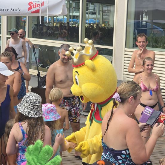 Eine Gruppe von Menschen, einschließlich Kindern, versammelt sich um einen Maskottchen, das in einem gelben Kostüm mit goldener Krone gekleidet ist. Das Maskottchen interagiert mit den Kindern, und einige Erwachsene stehen in der Nähe. Die Umgebung scheint ein Outdoor-Event zu sein, möglicherweise ein Festival.