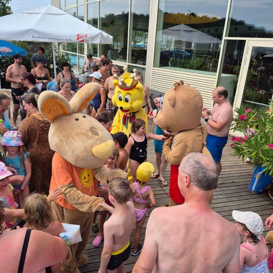 Eine Gruppe von Menschen, einschließlich Kindern, versammelt sich um zwei Maskottchen, eines als Bär und das andere als gelber Bär, bei einer Veranstaltung im Freien. Der Schauplatz scheint ein Resort oder Freizeitpark zu sein, mit einem Gebäude im Hintergrund.