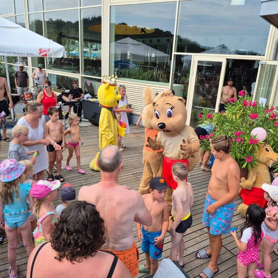 Eine Gruppe von Menschen, einschließlich Kindern, versammelt sich um einen Maskottchen, das als Bär verkleidet ist, auf einer Holzterrasse. Das Maskottchen trägt eine Krone und einen roten Rock. Hinter dem Maskottchen befinden sich Pflanzen mit rosa Blüten und ein Gebäude mit Glaswänden.