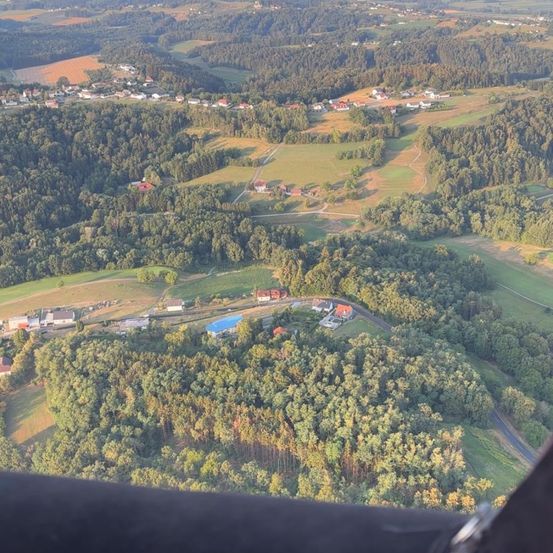 Eine Luftaufnahme einer ländlichen Landschaft mit dichten grünen Wäldern, verstreuten Häusern und einem blauen Swimmingpool in der Mitte.
