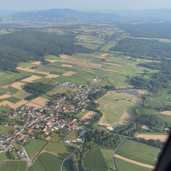 Luftaufnahme eines kleinen Dorfes, umgeben von üppigen grünen Feldern und Wäldern, mit Bergen in der Ferne.