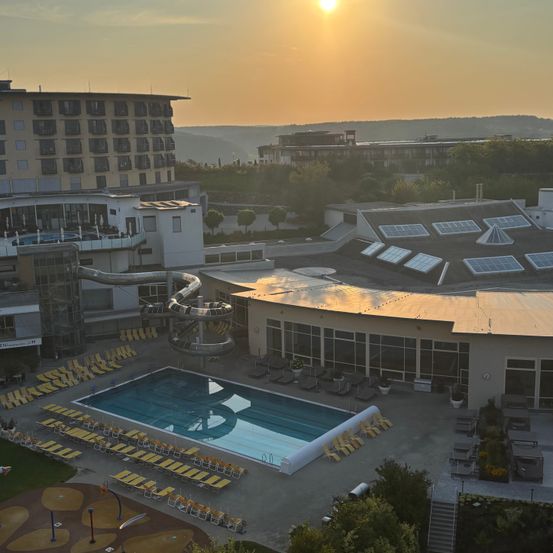 Ein Blick aus der Luft auf ein Resort mit einem großen Schwimmbad, Sonnenliegen und einer Wasserrutsche. Die Sonne geht im Hintergrund unter.