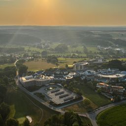 Eine Luftaufnahme eines modernen Architekturkomplexes in einer üppigen, grünen Landschaft mit einer untergehenden Sonne, die ein warmes Leuchten über die Szene wirft.