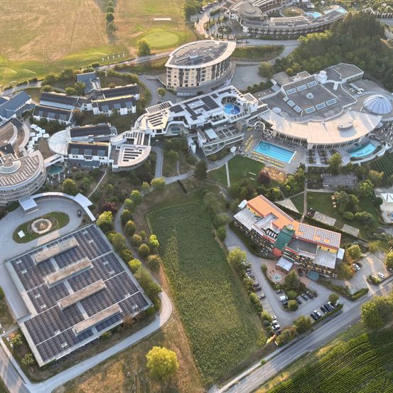 Eine Luftaufnahme eines modernen architektonischen Komplexes mit Solarpanelen, einem Schwimmbad und einem Golfplatz. Gebäude sind von grünen Feldern und Bäumen umgeben.