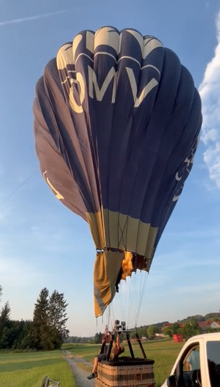 Ein Heißluftballon mit den Buchstaben OMV an der Seite, der von einem Van auf einem Grasfeld mit Bäumen im Hintergrund gezogen wird.