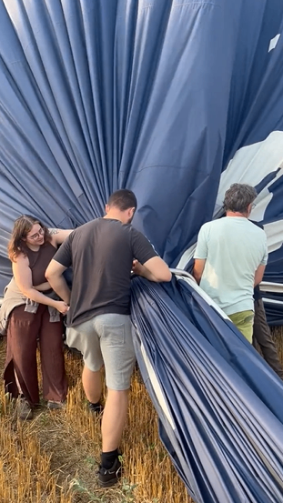 Drei Personen arbeiten gemeinsam daran, einen blauen Heißluftballon auf einem Feld aufzublasen.