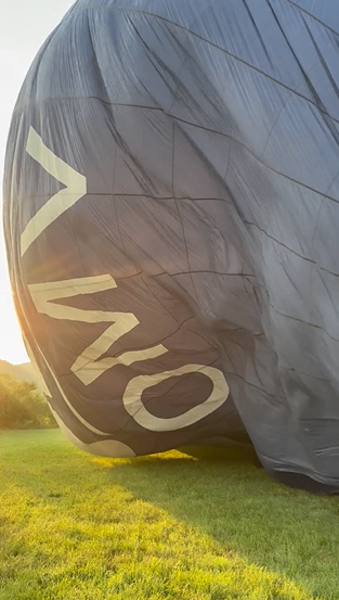 Ein Heißluftballon mit dem Wort 'VIO' auf der Seite, entleert und auf einem Grasfeld liegend, mit der untergehenden Sonne im Hintergrund.