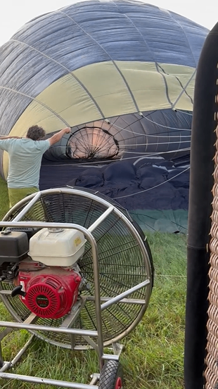 Eine Person steht neben einem Heißluftballon und hält dessen Stoff fest, während ein Ventilator am Ballon befestigt ist.