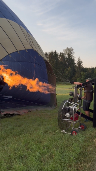 Ein Heißluftballon mit Flammen aus seinem Brenner wird auf einem Rasenfeld mit Bäumen im Hintergrund für den Start vorbereitet.