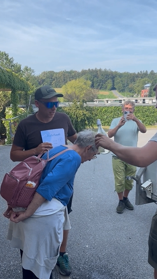 Eine Gruppe von Menschen versammelt sich auf einer Straße. Eine ältere Frau beugt sich vor und hält ein Papier in der Hand, während eine andere Person ihr Wasser auf den Kopf gießt. Hinter ihnen macht ein Mann ein Foto mit seinem Handy.
