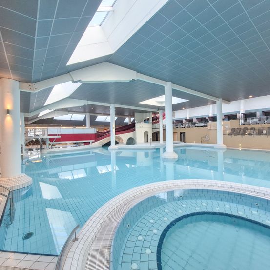 Ein Hallenbad mit blauer Wasseroberfläche, einer weißen Säule und einer roten Rutsche. Die Decke hat Lichter und ein Oberlicht.