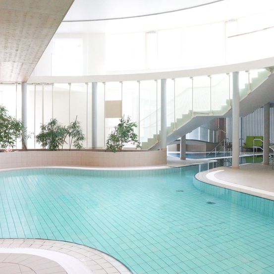 Ein runder Innenpool mit blauen Fliesen und einer Glastreppe im Hintergrund. Der Pool ist leer mit einer Reflexion der Treppe. Es gibt Topfpflanzen auf der linken Seite.