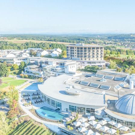 Luftaufnahme eines modernen Resorts mit Gebäuden, einem Pool und grünen Landschaften. Bäume umgeben das Gebiet. Berge sind in der Ferne.