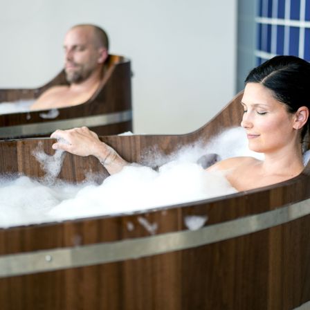 Ein Mann und eine Frau befinden sich in hölzernen Wannen voller Schaum, wirken entspannt. Der Raum hat eine blaue Wand.