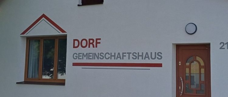 Ein weißes Gebäude mit einer braunen Tür und dem Schild Dorf Gemeinschaftshaus. Draußen steht eine Holzbank und ein Holztisch. Die Nummer 21 ist an der Wand angebracht.