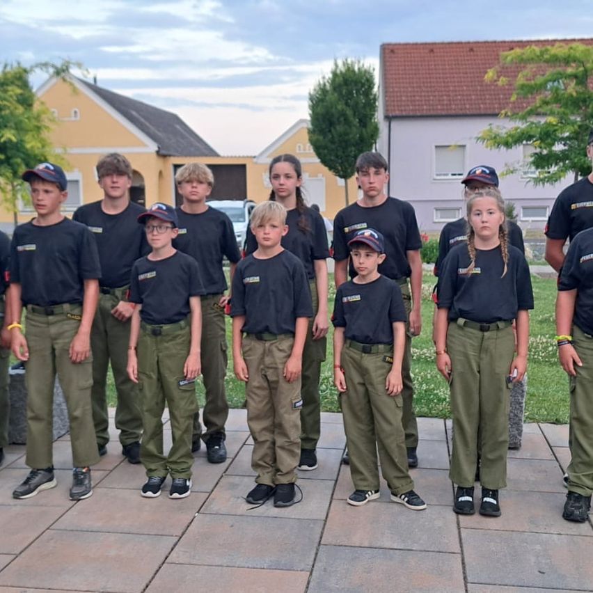 Eine Gruppe von Kindern in einheitlichen Outfits steht vor einem Haus mit Bäumen und einem bewölkten Himmel im Hintergrund.
