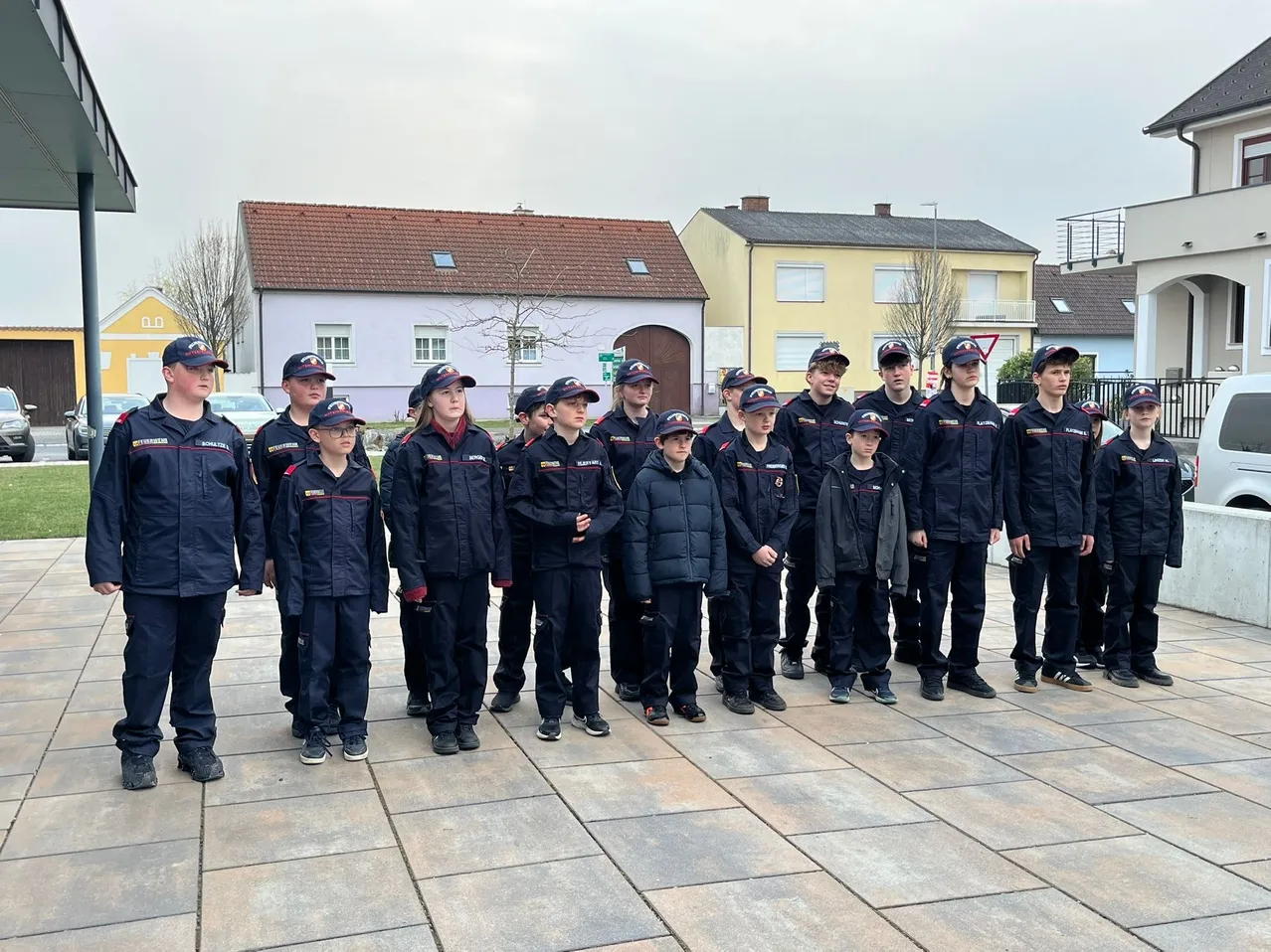 Eine Gruppe junger Menschen in Uniformen steht in einer Reihe im Freien mit Häusern und Bäumen im Hintergrund.
