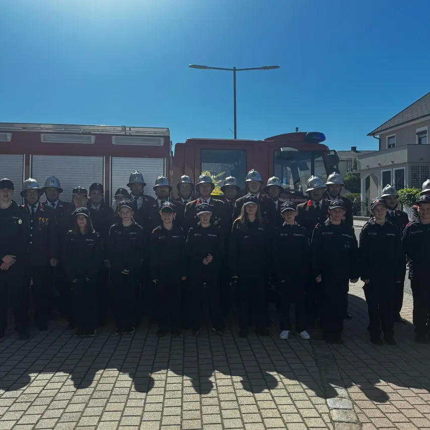 Eine Gruppe von Feuerwehrleuten in schwarzen Uniformen posiert für ein Foto vor einem Feuerwehrauto an einem sonnigen Tag.