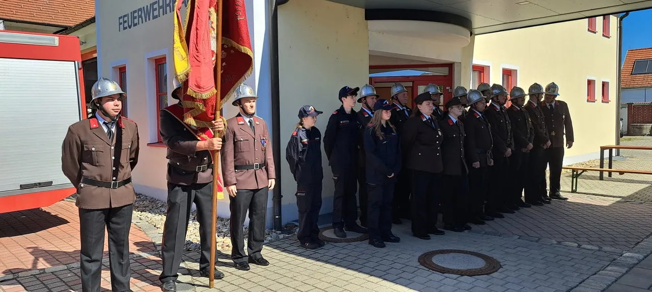 Eine Gruppe von Feuerwehrleuten in Uniform steht vor einem Gebäude, hält eine Fahne und trägt Helme. Ein Mann vorne hält einen Fahnenmast.