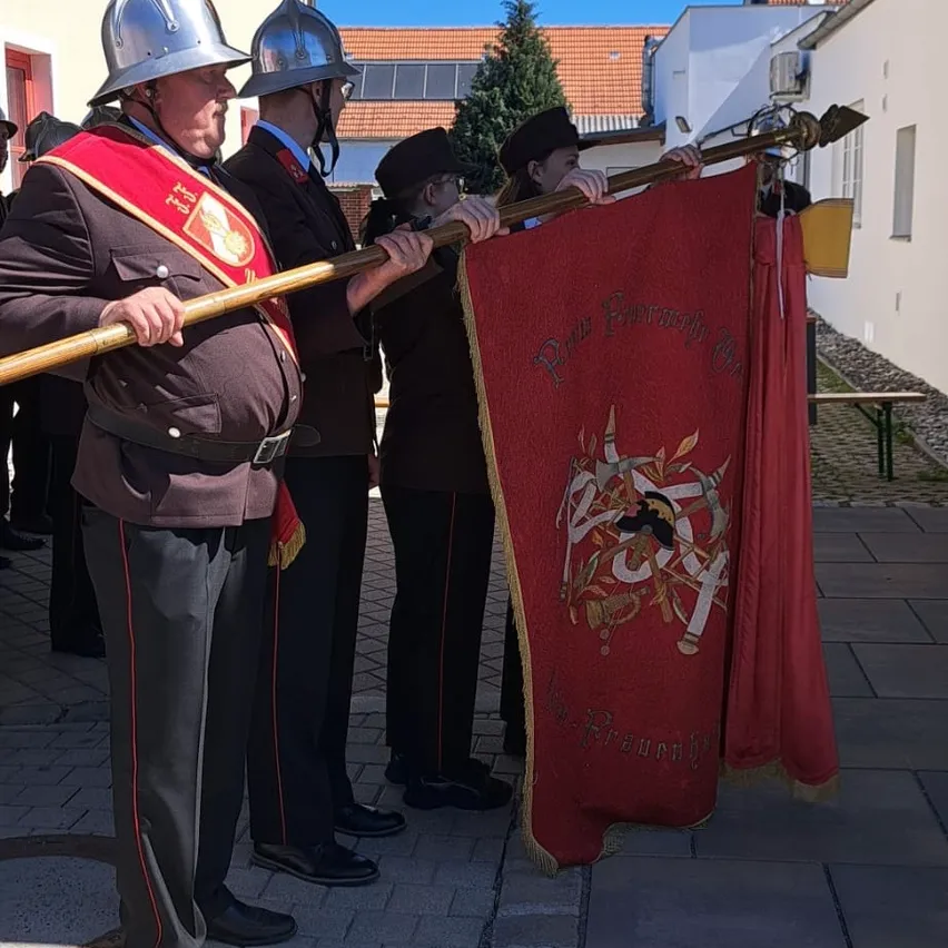 Eine Gruppe von Menschen in Uniformen, einschließlich eines Mannes, der eine rote Flagge mit goldenem Design hält, stehen auf einer gepflasterten Fläche. Dahinter befindet sich ein Gebäude mit einem Weihnachtsbaum und einer Bank.