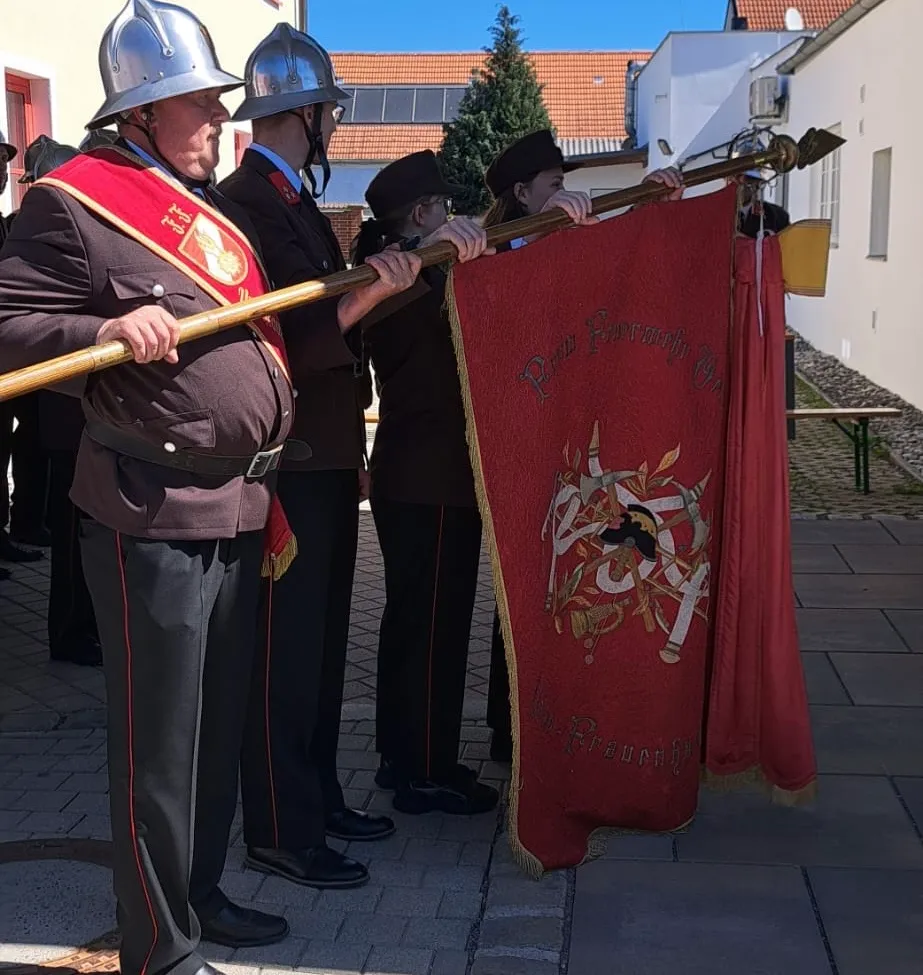 Eine Gruppe von Menschen in Uniformen, einschließlich eines Mannes, der eine rote Flagge mit goldenem Design hält, stehen auf einer gepflasterten Fläche. Dahinter befindet sich ein Gebäude mit einem Weihnachtsbaum und einer Bank.