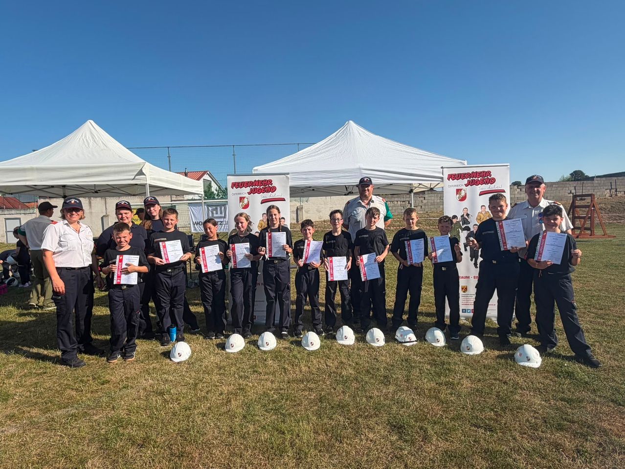A group of young people in black uniforms and white hats are posing for a photo. They are holding certificates. Behind them are two tents with banners. In the distance are buildings and a clear blue sky.