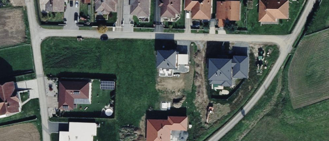 Luftaufnahme eines Wohngebiets mit verschiedenen Häusern und einer Straße. Einige Häuser haben Solarpanels auf ihren Dächern. Grüne Rasenflächen und Bäume sind sichtbar, ebenso wie ein geparktes Fahrzeug in der Nähe.