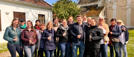 Eine Gruppe von Menschen steht auf einer Rasenfläche, hält Weingläser in der Hand und posiert für ein Foto. Sie tragen Winterkleidung, und einige tragen Brillen. Sie stehen vor einem Gebäude mit weißer Wand und braunem Dach. Dahinter befinden sich Bäume und ein weiteres Gebäude.