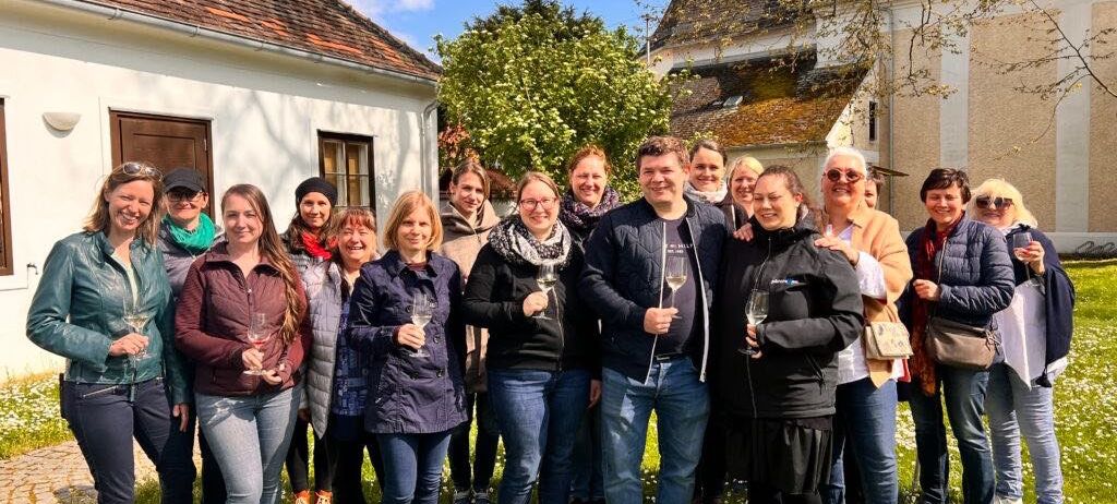 Eine Gruppe von Menschen steht auf einer Rasenfläche, hält Weingläser in der Hand und posiert für ein Foto. Sie tragen Winterkleidung, und einige tragen Brillen. Sie stehen vor einem Gebäude mit weißer Wand und braunem Dach. Dahinter befinden sich Bäume und ein weiteres Gebäude.