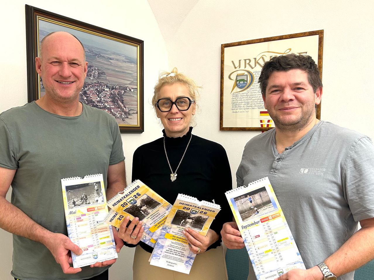Drei Personen halten Kalender in die Höhe und lächeln. Die Frau trägt eine Brille und eine Halskette. Hinter ihnen hängen zwei gerahmte Bilder an der Wand. Das linke Bild zeigt eine Stadtlandschaft und das rechte ein Wappen mit Text.