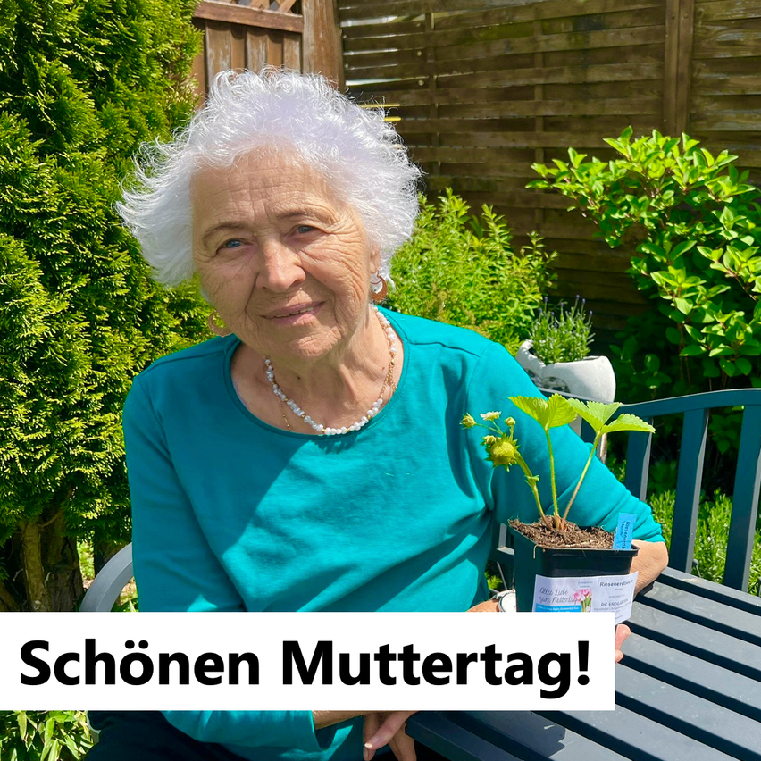 An elderly woman in a garden, smiling, holds a potted plant. She wears a necklace and a teal top. The image has a text overlay that says 'Schönen Muttertag!' in German.